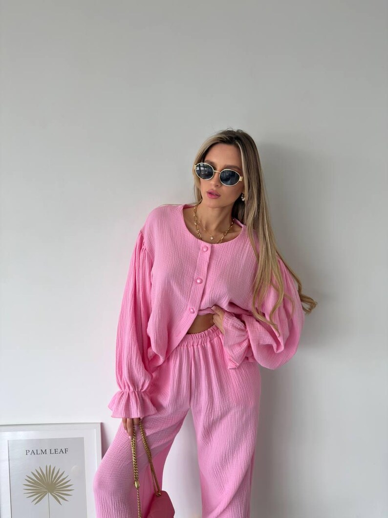 May include: A bright pink two-piece set consisting of a long-sleeved button-down shirt with ruffled cuffs and matching high-waisted trousers. The fabric has a textured, crinkled appearance. The shirt features a round neckline and three buttons.