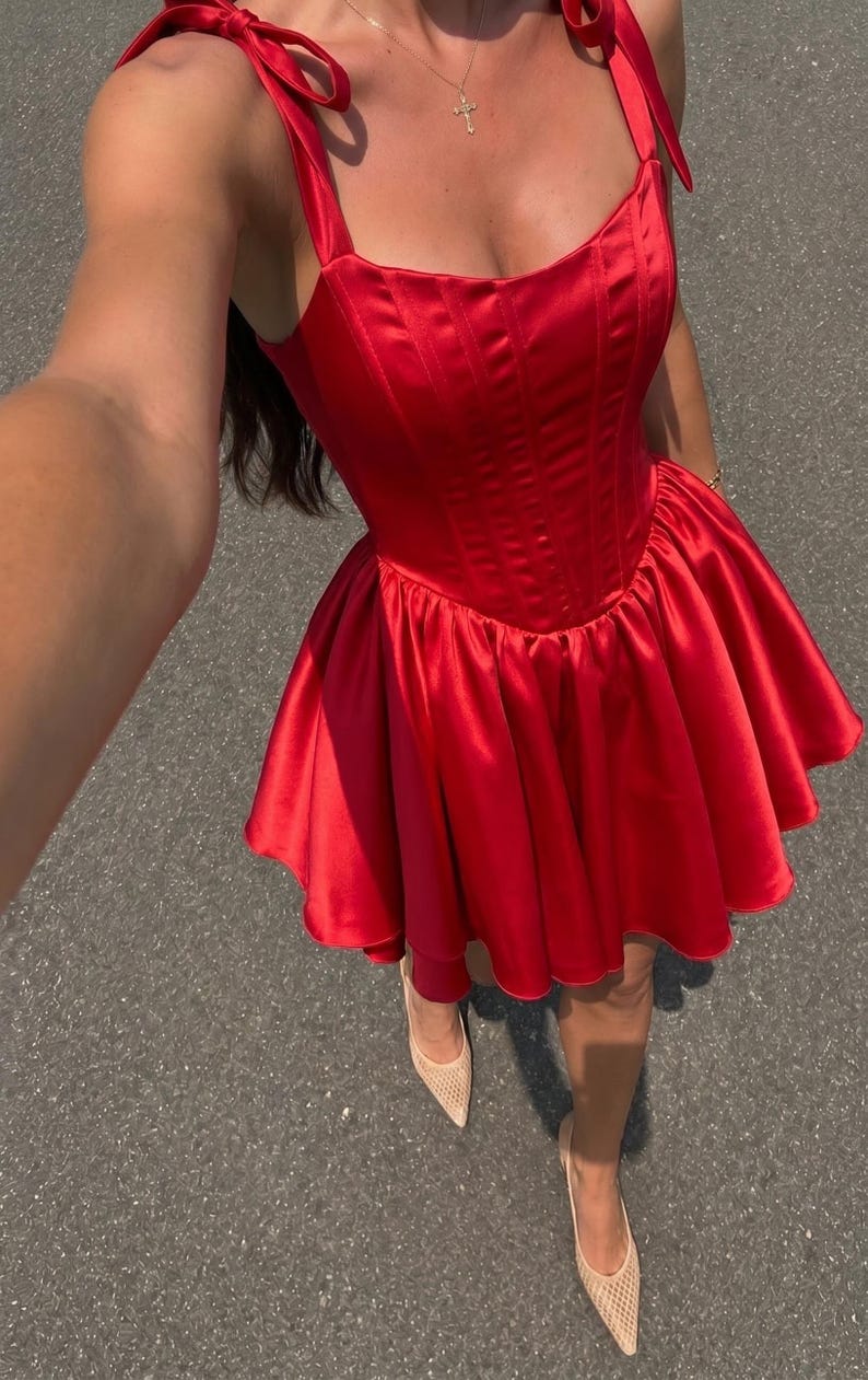 May include: A vibrant red satin mini dress featuring a structured corset-style bodice with vertical seams, a square neckline, and shoulder straps that tie into bows. The skirt is gathered at the waist, creating a flared, voluminous silhouette.