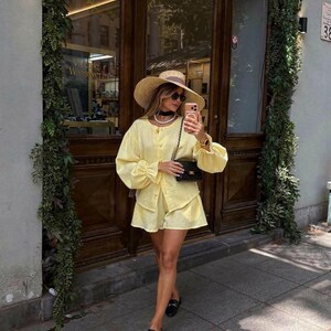 May include: A light yellow long-sleeved button-down shirt and matching shorts set, paired with a wide-brimmed straw hat, a black quilted shoulder bag, a pearl necklace, a black choker, and black loafers with gold-tone hardware details.