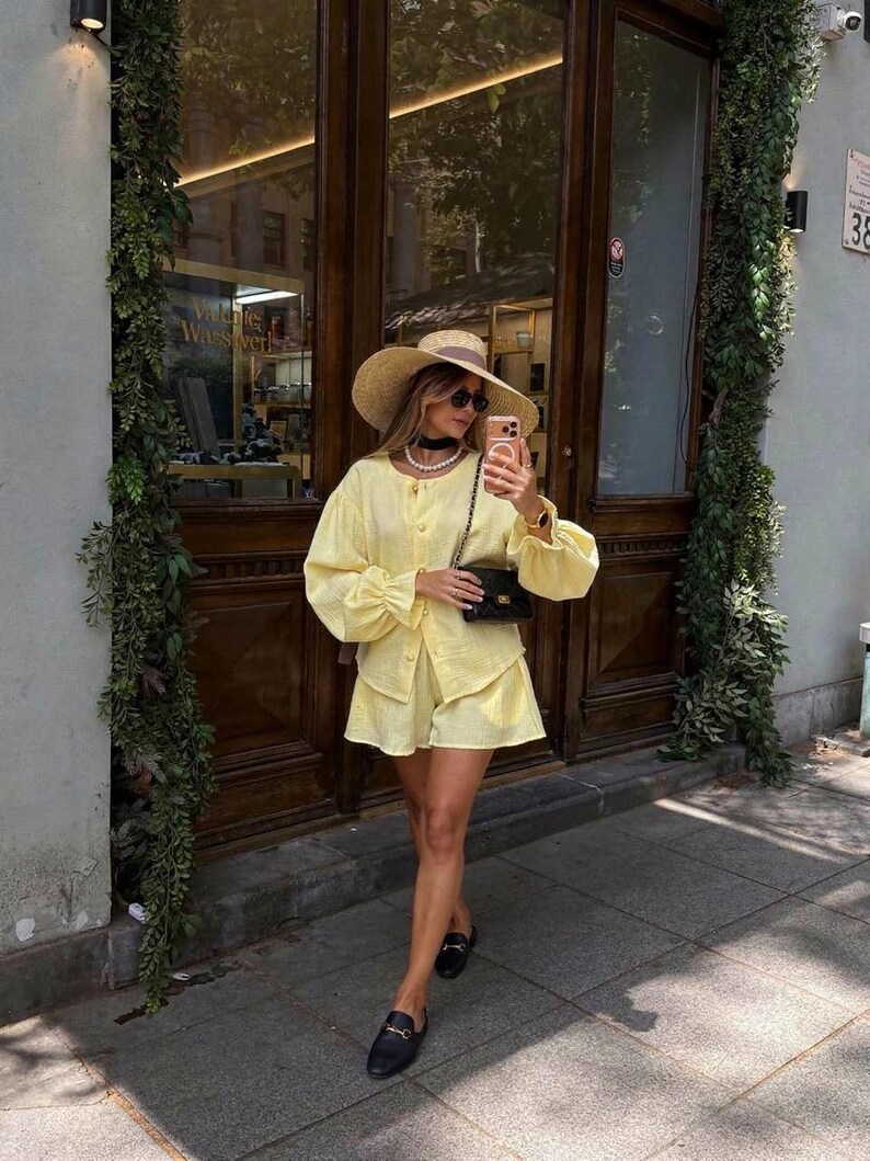 May include: A light yellow long-sleeved button-down shirt and matching shorts set, paired with a wide-brimmed straw hat, a black quilted shoulder bag, a pearl necklace, a black choker, and black loafers with gold-tone hardware details.
