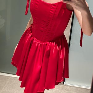 May include: A vibrant red satin mini dress featuring a structured corset-style bodice with vertical seams and a flared, pleated skirt. The garment includes thin shoulder straps that tie at the top, creating a sleeveless silhouette with a smooth finish.