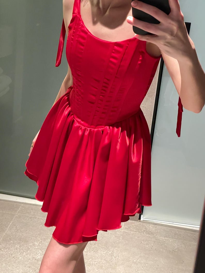 May include: A vibrant red satin mini dress featuring a structured corset-style bodice with vertical seams and a flared, pleated skirt. The garment includes thin shoulder straps that tie at the top, creating a sleeveless silhouette with a smooth finish.
