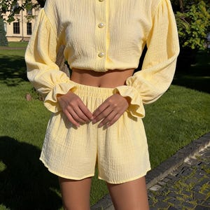May include: A light yellow two-piece set consisting of a long-sleeved cropped blouse and matching shorts. The blouse features a round neckline, three pearl-like buttons, and gathered sleeves with ruffled cuffs. The shorts have an elastic waistband.