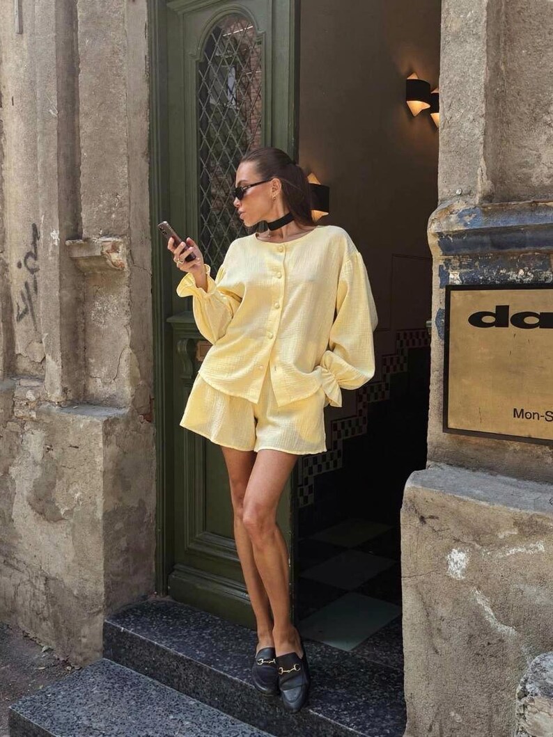 May include: A light yellow two-piece set consisting of a long-sleeved button-down shirt with gathered cuffs and matching loose-fitting shorts. The fabric has a subtle textured finish. The outfit is paired with black loafers and a black choker.