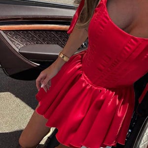 May include: A vibrant red satin mini dress featuring a structured corset-style bodice with vertical stitching, a square neckline, and wide shoulder straps. The skirt is gathered at the waist, creating a voluminous, flared silhouette with a smooth finish.