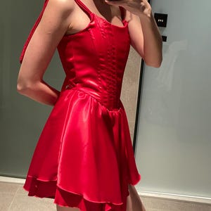 May include: A vibrant red satin mini dress featuring a structured corset-style bodice with vertical stitching, a square neckline, and wide shoulder straps that tie at the top. The skirt is flared with a layered, ruffled hemline in a smooth finish.