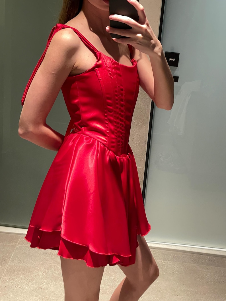 May include: A vibrant red satin mini dress featuring a structured corset-style bodice with vertical stitching, a square neckline, and wide shoulder straps that tie at the top. The skirt is flared with a layered, ruffled hemline in a smooth finish.