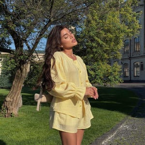 May include: A light yellow two-piece set consisting of a long-sleeved button-down shirt with gathered cuffs and matching shorts. The fabric has a textured, crinkled appearance. A small beige shoulder bag is visible hanging from the shoulder.