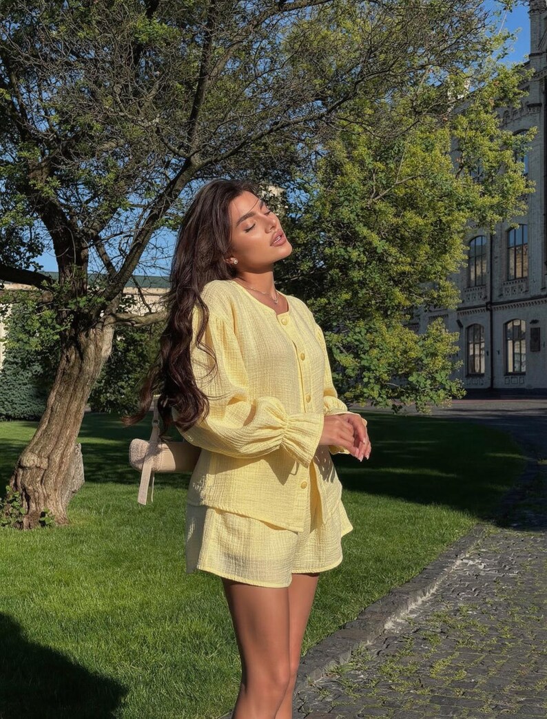 May include: A light yellow two-piece set consisting of a long-sleeved button-down shirt with gathered cuffs and matching shorts. The fabric has a textured, crinkled appearance. A small beige shoulder bag is visible hanging from the shoulder.