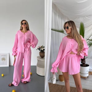 May include: A two-piece set consisting of a light pink, long-sleeved button-down shirt with gathered cuffs and matching wide-leg trousers or shorts, both crafted from a textured, crinkled fabric with a round neckline and front button closure.