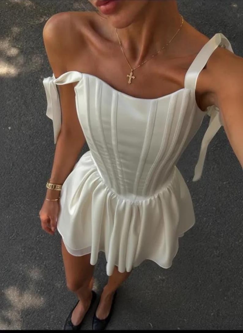 May include: A white sleeveless mini dress featuring a structured corset-style bodice with vertical boning, a flared skirt, and shoulder straps with tie-up details. The garment is made of a smooth, satin-like fabric with a subtle sheen throughout.