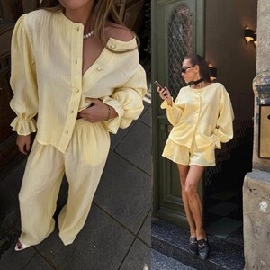 May include: A light yellow two-piece set consisting of a long-sleeved button-down shirt with gathered cuffs and matching wide-leg trousers. The shirt features a round neckline and pearl-like buttons, while the trousers have a relaxed fit.