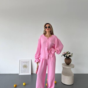 May include: A bright pink two-piece set consisting of a long-sleeved button-down shirt with ruffled cuffs and matching wide-leg trousers. The fabric has a subtle crinkled texture. The outfit is paired with dark brown sandals and gold jewelry.