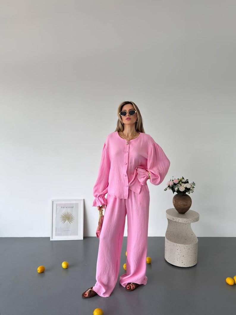 May include: A bright pink two-piece set consisting of a long-sleeved button-down shirt with ruffled cuffs and matching wide-leg trousers. The fabric has a subtle crinkled texture. The outfit is paired with dark brown sandals and gold jewelry.