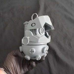 Może przedstawiać: Szary, drukowany w 3D obiekt o mechanicznym wyglądzie z zawiasową górą przypominającą stylizowaną głowę zwierzęcia. Obiekt posiada okrągły otwór z szczegółowym wnętrzem, detale w kształcie plusa i uchwyt w kształcie pierścienia.