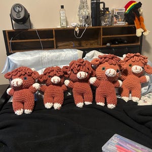 May include: Five handmade crochet Highland cow plushies. Each plushie features a reddish-brown body, a fluffy head, and white accents on the face and hooves. The cows are arranged in a row, showcasing their identical design and craftsmanship.