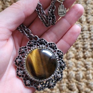Op de afbeelding: Een handgemaakte ketting met een grote, ronde tijgeroogsteen gezet in een sierlijke, filigraan-stijl zilverkleurige hanger. De hanger hangt aan een zilveren ketting. Een kleine bedel met de woorden "Hand Made" is bevestigd.