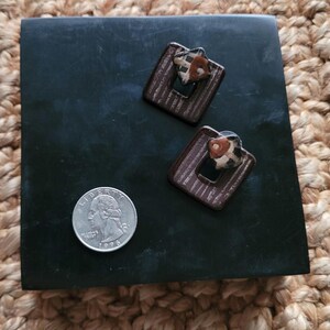 May include: A pair of brown and white ceramic earrings with a geometric design. The earrings have a square shape with a smaller square cut out in the center. The earrings are decorated with a brown and white striped pattern and a small brown and white patterned circle in the center.