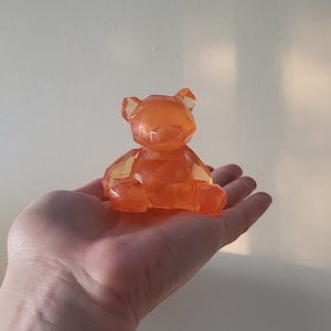 Könnte beinhalten: Eine kleine, orangefarbene, geometrische Bärenfigur aus Harz. Der Bär sitzt mit gekreuzten Beinen und hat ein schlichtes, stilisiertes Design.