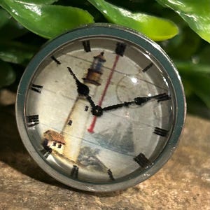 May include: A silver-toned ring with a clear, domed face featuring a lighthouse scene. The clock face has black Roman numerals and black hands. The background includes a red line and a blue and white seascape.