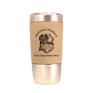 May include: A stainless steel travel mug with a tan faux leather wrap. The wrap features a black and white line drawing of an Australian Shepherd dog's head with the text "Australian Shepherd" above the dog and "Your Dogs Name Here" below the dog.