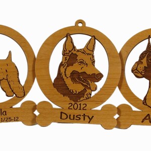 May include: Three wooden dog ornaments with engraved portraits of dogs and their names. The ornaments are attached to a bone-shaped piece with the text "Bella 4/10/02 - 1/25-12", "2012 Dusty", and "Ace".
