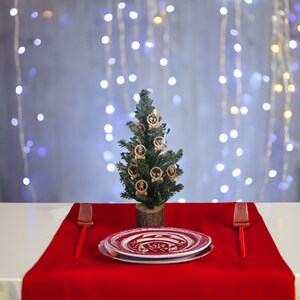 May include: A festive holiday table setting featuring a small Christmas tree adorned with wooden ornaments. A red tablecloth is set with plates and red serving utensils. The background has a string of white lights.