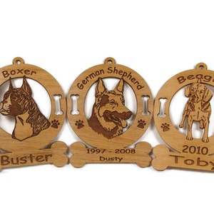 K&ouml;nnte beinhalten: Drei h&ouml;lzerne Hundefiguren als Weihnachtsbaumschmuck mit gravierten Bildern eines Boxers, eines Deutschen Sch&auml;ferhundes und eines Beagles. Jeder Anh&auml;nger hat ein knochenf&ouml;rmiges Schild mit dem Namen des Hundes und den Daten. Der Boxer-Anh&auml;nger sagt "Buster", der Deutsche Sch&auml;ferhund-Anh&auml;nger sagt "Dusty 1997-2008" und der Beagle-Anh&auml;nger sagt "Toby 2010".