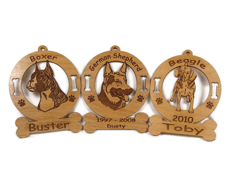 K&ouml;nnte beinhalten: Drei h&ouml;lzerne Hundefiguren als Weihnachtsbaumschmuck mit gravierten Bildern eines Boxers, eines Deutschen Sch&auml;ferhundes und eines Beagles. Jeder Anh&auml;nger hat ein knochenf&ouml;rmiges Schild mit dem Namen des Hundes und den Daten. Der Boxer-Anh&auml;nger sagt "Buster", der Deutsche Sch&auml;ferhund-Anh&auml;nger sagt "Dusty 1997-2008" und der Beagle-Anh&auml;nger sagt "Toby 2010".