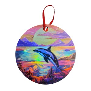 May include: Round Christmas ornament featuring a vibrant ocean scene with a leaping dolphin. The ornament has a red ribbon for hanging. The artwork includes a blue and white dolphin, mountains, and a colorful sunset.