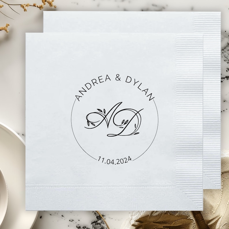 May include: White paper napkin with a black circle design featuring the text "Andrea & Dylan" and the initials "AD" with floral accents. The date "11.04.2024" is printed below the circle.