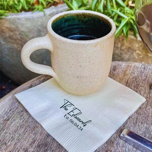 May include: A white ceramic mug with a green interior and a black handle sits on a white napkin with the text "The Edwards Est. 06.04.24" printed on it.