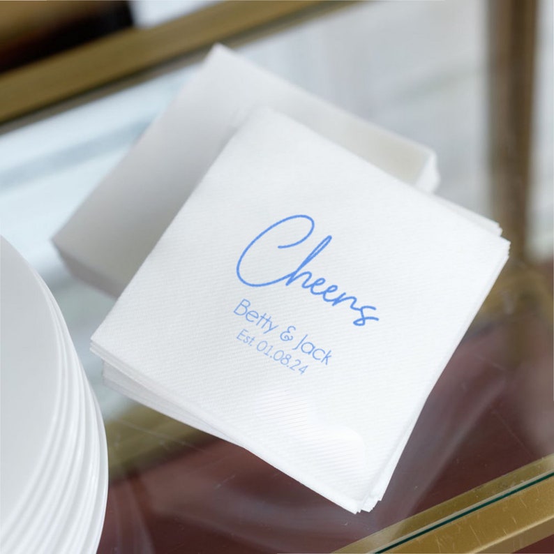 May include: White paper napkins with blue text that reads "Cheers" and "Betty & Jack Est 01.08.24".