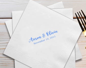 May include: White paper napkins with blue text reading "Aaron & Olivia" and "November 10, 2023". The napkins are stacked on a light wood surface, with a gold and black fork in the background. The napkins have a textured border.
