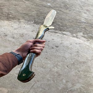 Op de afbeelding: Een lang mes met een zwart en groene handvat en een zilveren lemmet. Het handvat heeft een decoratief ontwerp en twee messing accenten.