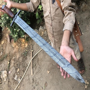 May include: A long, silver sword with a brown leather handle. The blade has a distinctive pattern of swirling lines, known as Damascus steel.