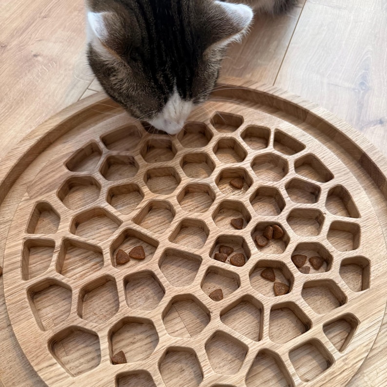 May include: A wooden cat puzzle feeder with a honeycomb design. The feeder is filled with cat food and a cat is shown sniffing the food. The feeder is made of light-colored wood and has a round shape.