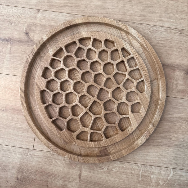 May include: A round, wooden serving tray with a honeycomb pattern carved into the center. The tray is made of light brown wood and has a raised edge. The tray is approximately 12 inches in diameter.