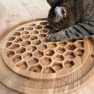 May include: A wooden cat food puzzle with a honeycomb design. The circular puzzle is made of light-colored wood and features numerous hexagonal cutouts. A tabby cat is partially visible, interacting with the puzzle.
