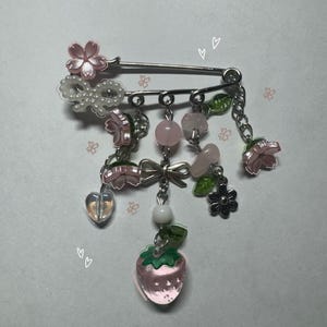 Puede incluir: Broche decorativo con base de imperdible plateado adornado con flores rosas, cuentas y un dije de fresa colgante. El broche incluye un lazo, un corazón y detalles florales, creando un accesorio caprichoso.