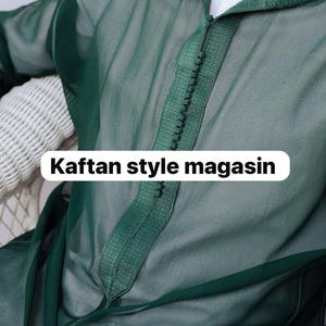 Può includere: Un caftano verde smeraldo trasparente con cappuccio e abbottonatura. Il caftano presenta un bordo decorativo e il testo "Kaftan style magasin" in un rettangolo bianco.
