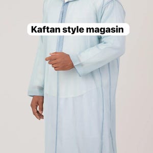 Może przedstawiać: Jasnoniebieska szata w stylu kaftanu z kapturem i długimi rękawami. Szata ma niebieskie detale szwów i tekst "Kaftan style magasin" w białym polu. Odzież wykonana jest z lekkiej tkaniny.