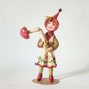 May include: A handmade doll with red hair and a red hat holding a mushroom. The doll is wearing a white dress with green and red floral embroidery. The doll is standing on a brown base.