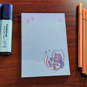 Könnte beinhalten: Eine Auswahl an Schreibwaren auf einer Holzoberfläche. Enthält drei Staedtler Textsurfer classic Textmarker in Pfirsich, Hellblau und Hellgrün, einen Notizblock mit einer Katzen-Astronauten-Illustration und drei farbige Stifte.