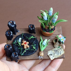Pode incluir: Um altar de madeira em miniatura com velas pretas, um homem de gengibre, uma planta em vaso com flores azuis, uma caveira com flores e um pedaço de pergaminho com um símbolo.