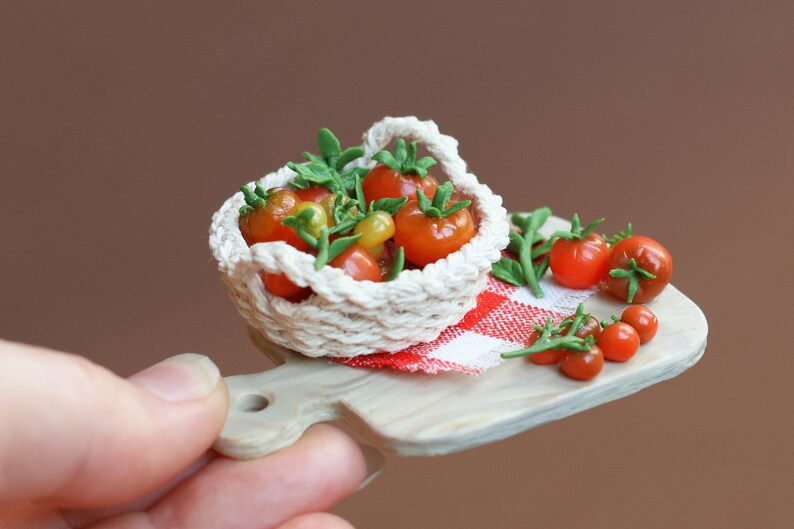 May include: A miniature white crocheted basket filled with red and yellow tomatoes, sitting on a wooden cutting board with a red and white checkered cloth. There are additional tomatoes scattered around the basket.