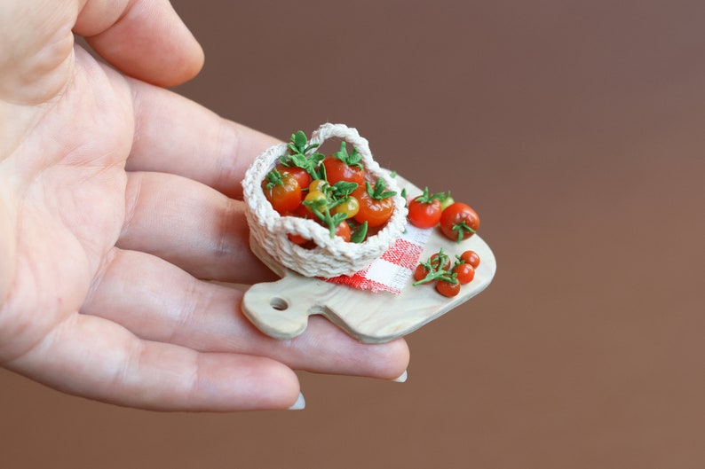 May include: A miniature wooden cutting board with a red and white checkered cloth, a small crocheted basket filled with red and yellow tomatoes, and additional tomatoes scattered around the basket.