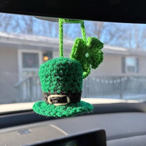 May include: A green crocheted leprechaun hat with a black belt and a green four-leaf clover hanging from the rearview mirror of a car.