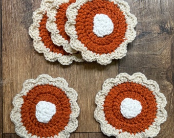 May include: A set of five circular crochet coasters featuring a central white circle, a larger burnt orange middle section, and a scalloped cream-colored border. The coasters are arranged with three stacked at the top and two placed side-by-side below.