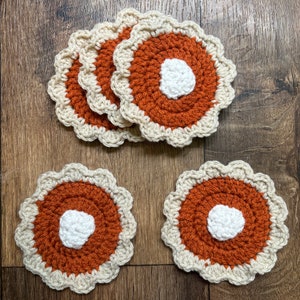 Peut inclure: Ensemble de quatre dessous-de-verre en crochet de couleur orange rouille avec un cercle central blanc. Les dessous-de-verre ont un bord festonné et sont faits d'un fil doux et texturé.